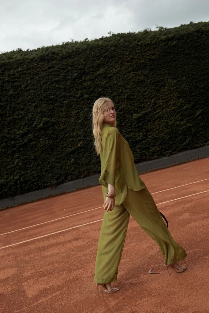 Side close-up of a blonde model in an olive green long set with resin bangles, holding a tennis racket near the net in a refined outdoor setting in Punta Cana.