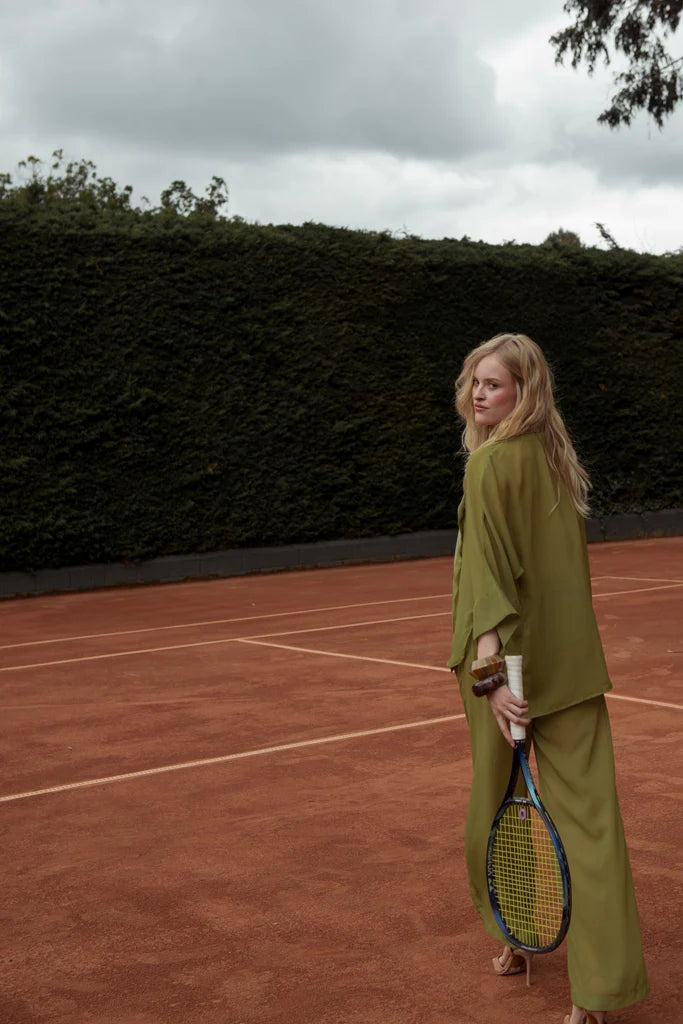 Model with loose blonde hair holding a tennis racket while posing from behind on a clay court in Punta Cana, dressed in a flowing olive green long set by ANT/BES, surrounded by lush tropical greenery.
