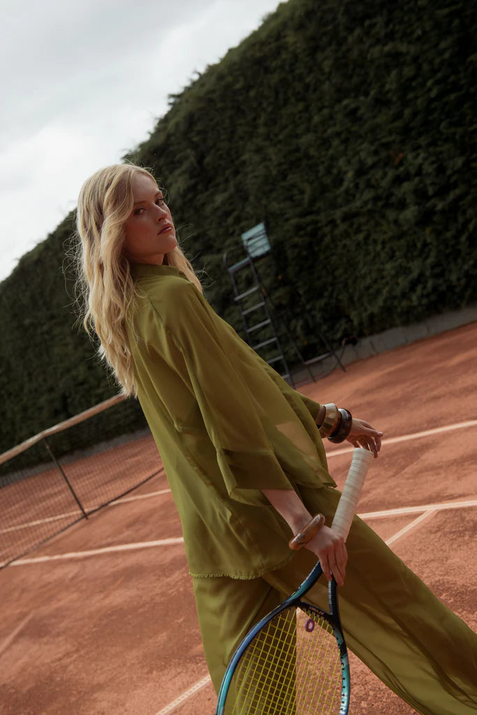 Blonde woman walking across a clay tennis court in Punta Cana wearing a sheer olive green long set from ANT/BES, paired with nude heels and an effortlessly elegant pose under a moody sky.