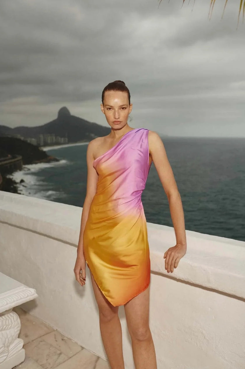 Model wearing the Celia Dress in Flamengo, showcasing its one-shoulder design and ombré gradient from pink to orange.