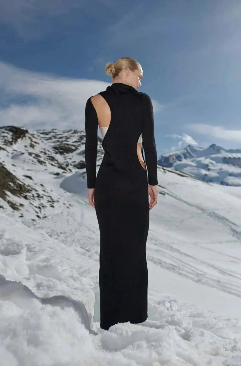 Back view of the Baobab Frida Dress in black with sculptural cutouts, set in a snowy mountain landscape.