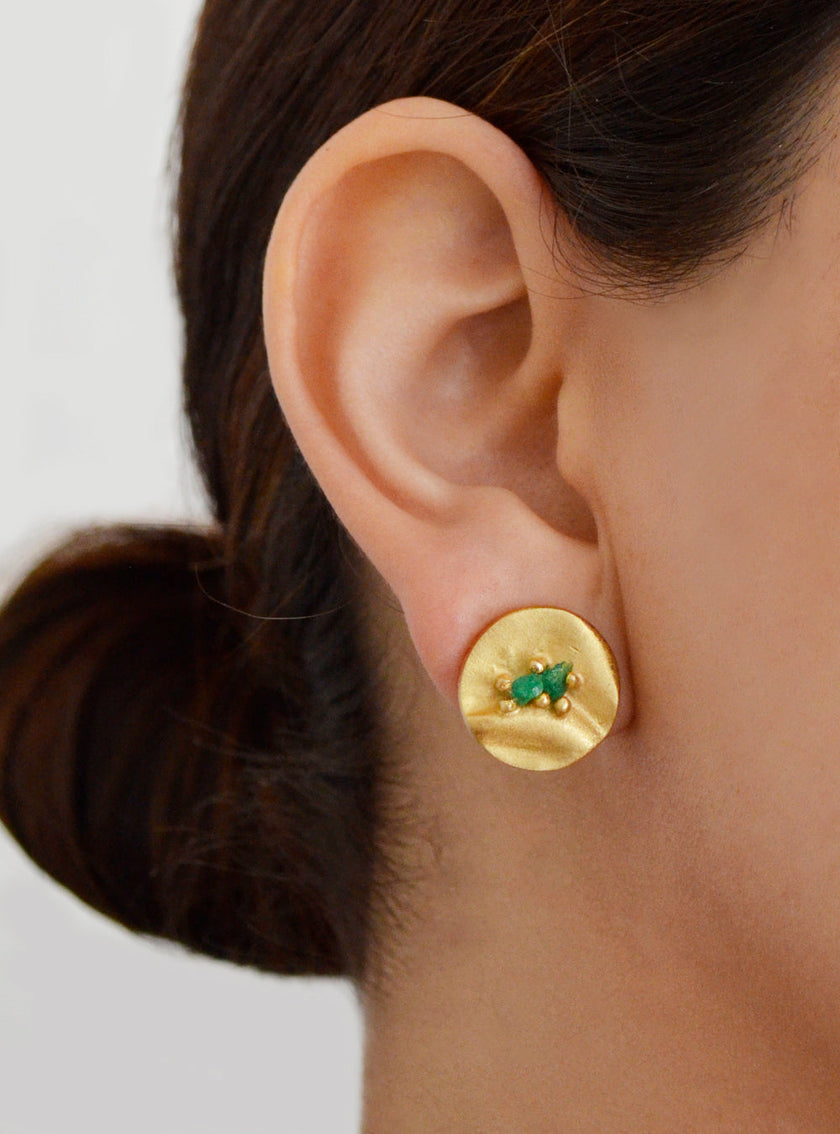 Close-up of a woman's ear wearing a round gold stud earring with an embedded Colombian emerald.