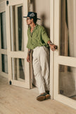 Man wearing a black Cross Charm hat and a green linen shirt, leaning against a wooden-framed glass door.