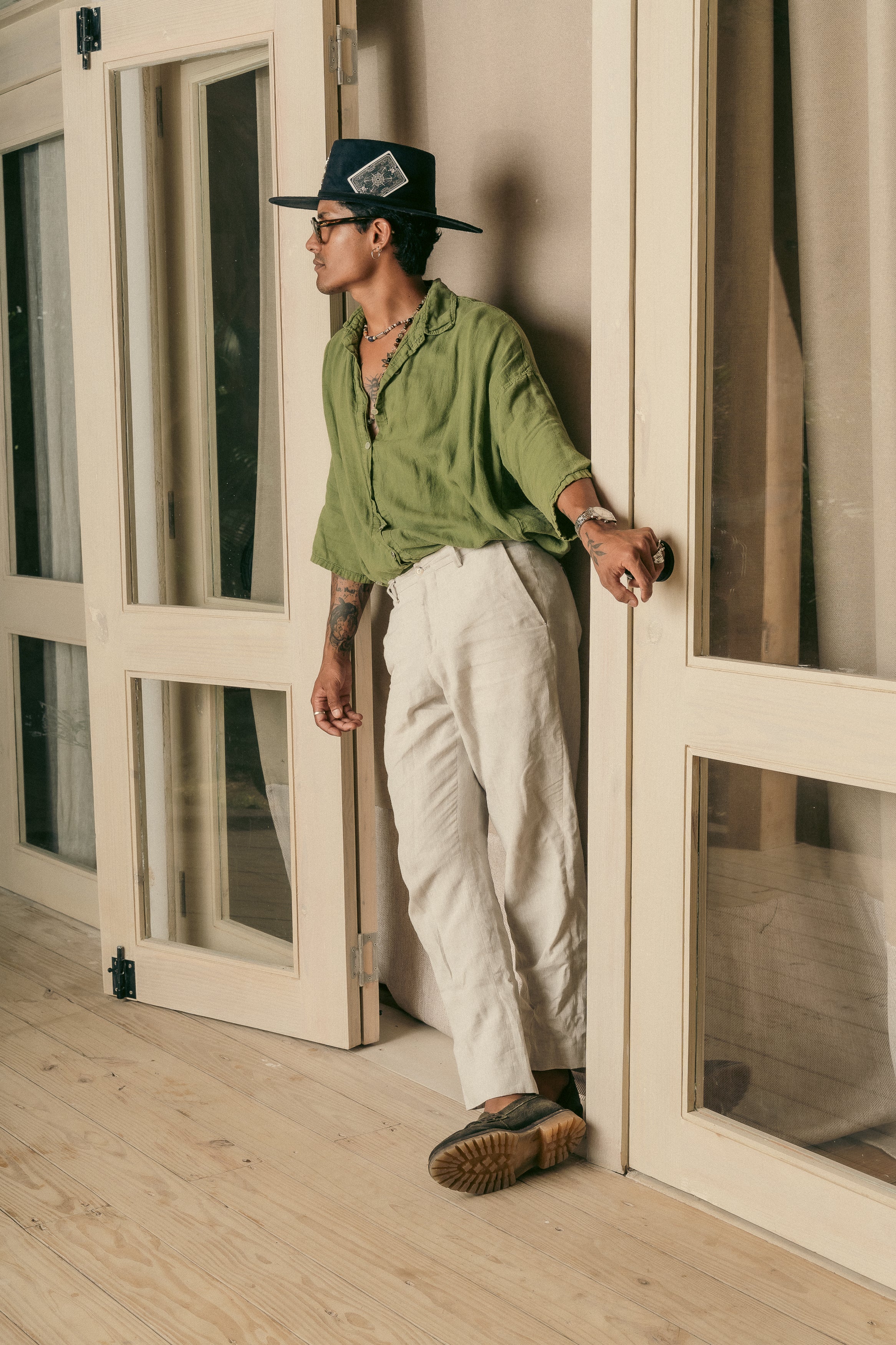 Man wearing a black Cross Charm hat and a green linen shirt, leaning against a wooden-framed glass door.