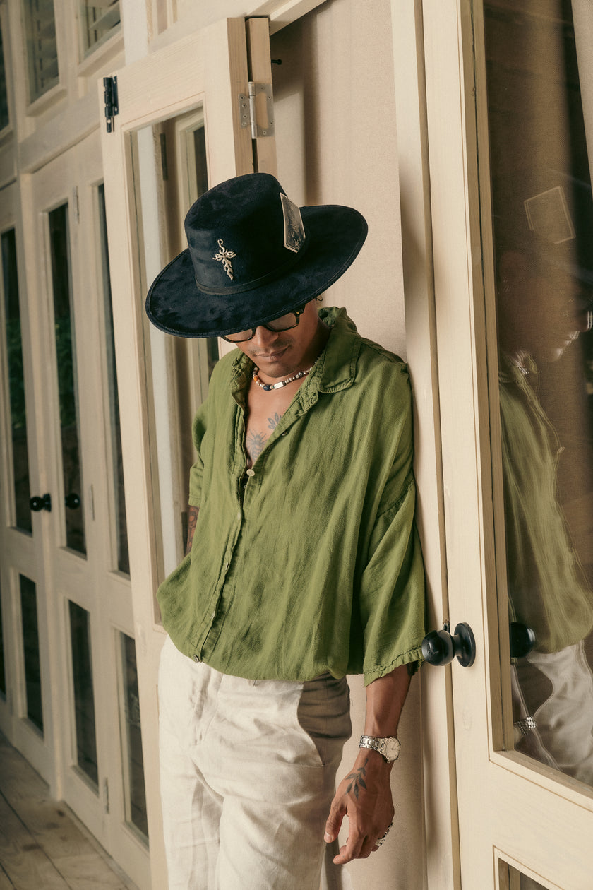 Close-up of a man wearing the black Cross Charm hat with flaming cross, standing against a mirror and wooden door