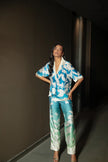 Smiling woman standing beside a large tropical plant in Punta Cana, dressed in the Eloisa Long Set by ANTIBES with a nature-inspired blue and green print, accessorized with a white designer handbag and sandals.