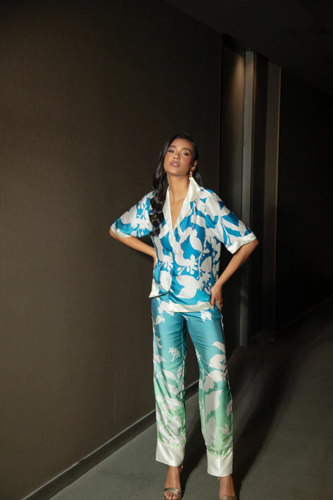 Smiling woman standing beside a large tropical plant in Punta Cana, dressed in the Eloisa Long Set by ANTIBES with a nature-inspired blue and green print, accessorized with a white designer handbag and sandals.