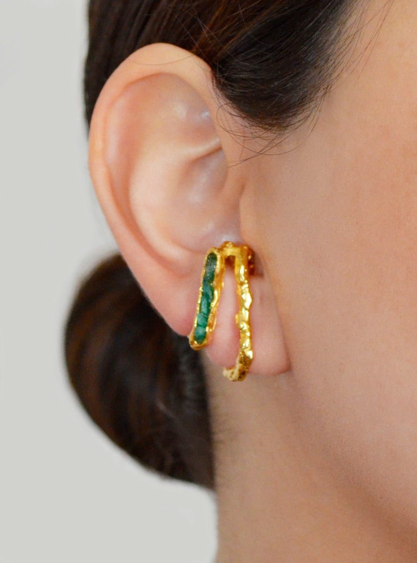 Close-up of a woman's ear adorned with a sculptural double hoop earring in gold with raw emeralds.