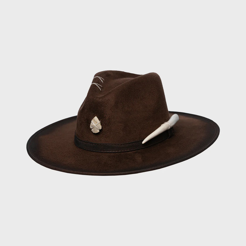 Side angle of brown artisan felt hat with cream talisman and hand-carved tooth accent, laid on light background.