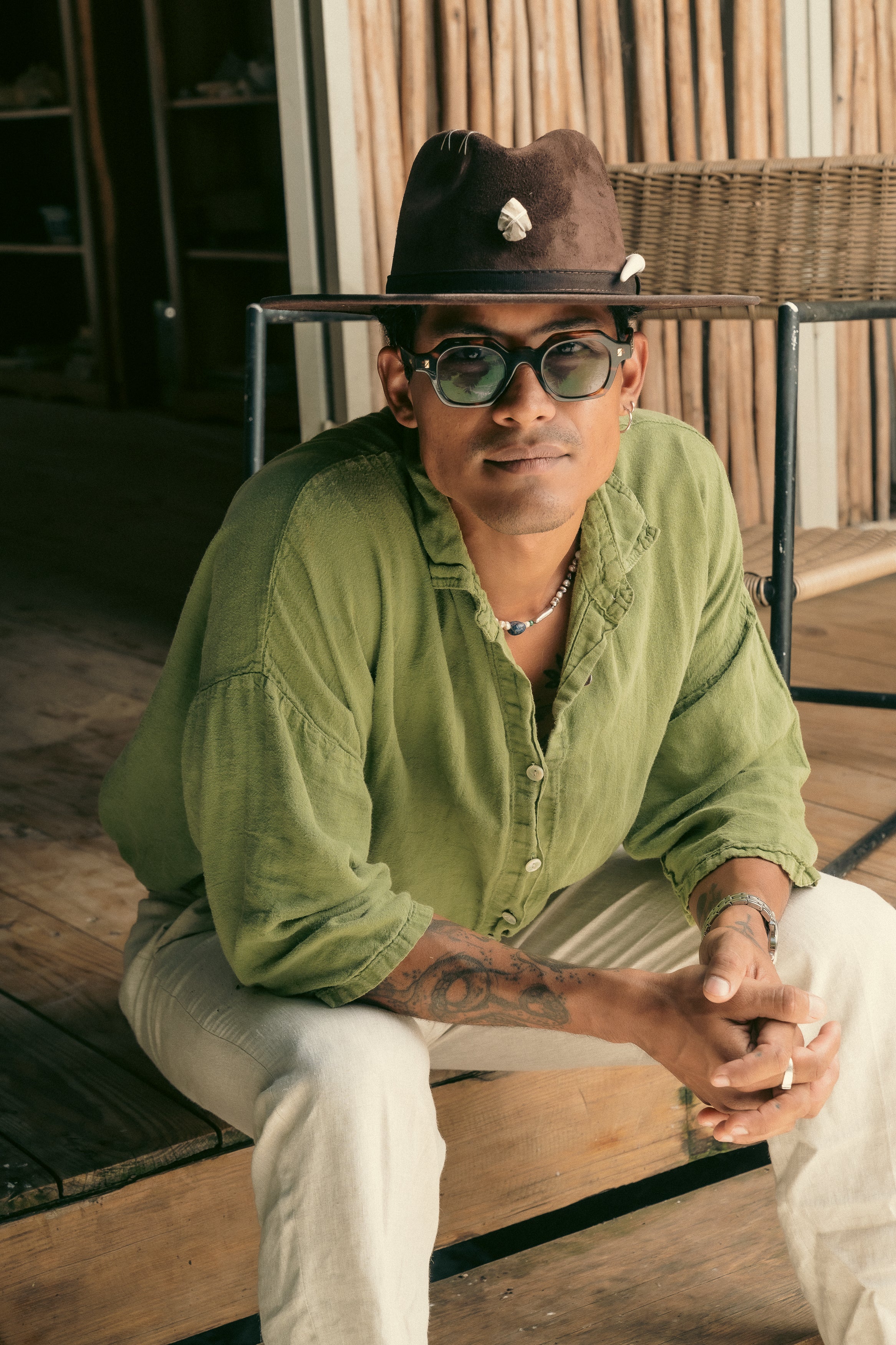Close-up of male model in sunglasses wearing brown handmade Hunter hat with ivory tribal accents, seated casually.