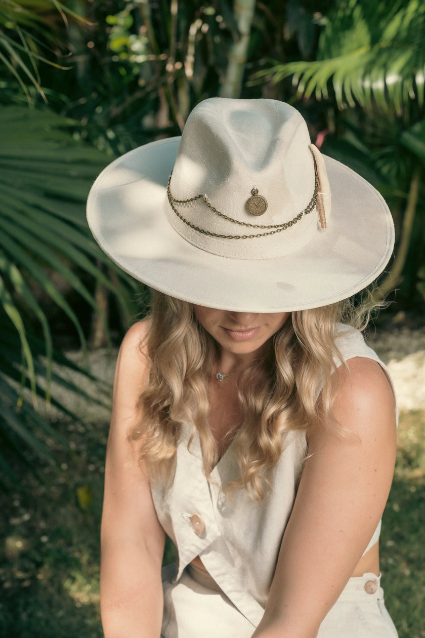 Blonde model in tropical garden wearing the Lucky Charm hat with wide brim and layered antique chains, exuding quiet elegance.