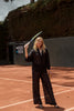 Close-up of the MAUI Long Set by ANTIBES in Punta Cana, highlighting the fluid black fabric and relaxed tailoring, styled with a tennis racket for an edgy, elevated sporty look.