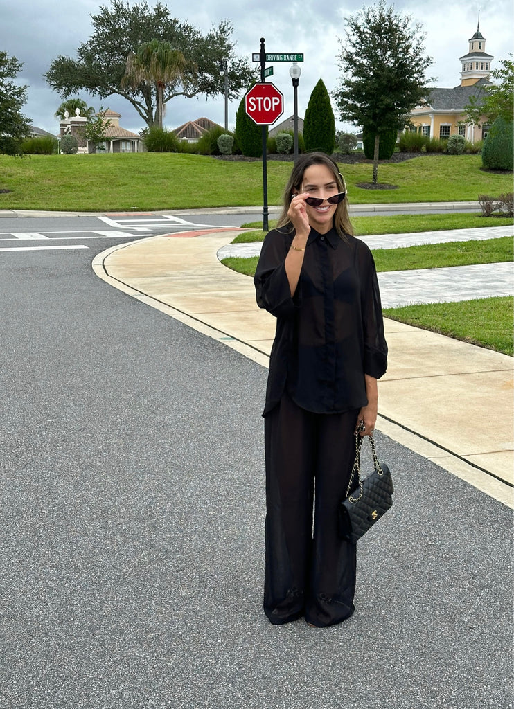 Model standing on a clay tennis court in Punta Cana, wearing the MAUI Long Set by ANTIBES — a sheer black ensemble paired with strappy heels and a tennis racket for a bold, fashion-forward statement.
