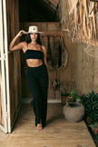 Full-body view of a model wearing the Mamba Charm hat, styled with a minimalist black ensemble, standing in a tropical wooden doorway with rustic accessories.