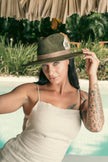 Woman with tattoos wearing the Olive Pirates hat and beige linen dress, under a palm-thatched roof near a tropical pool.