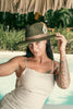 Woman with tattoos wearing the Olive Pirates hat and beige linen dress, under a palm-thatched roof near a tropical pool.