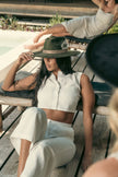 Model lounging on a wooden poolside deck chair, wearing the Olive Pirates hat with white linen outfit, evoking effortless summer elegance.