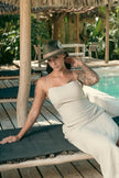 Model in beige dress wearing Olive Pirates hat while seated beneath tropical shade, merging relaxed island lifestyle with editorial flair.