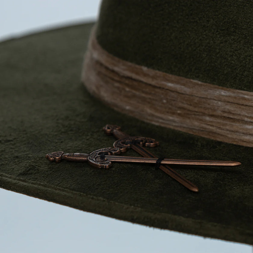 Macro shot of antique-style crossed sword pin on the brim of the Olive Pirates hat, highlighting aged bronze finish and craftsmanship.