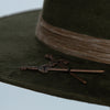 Macro shot of antique-style crossed sword pin on the brim of the Olive Pirates hat, highlighting aged bronze finish and craftsmanship.
