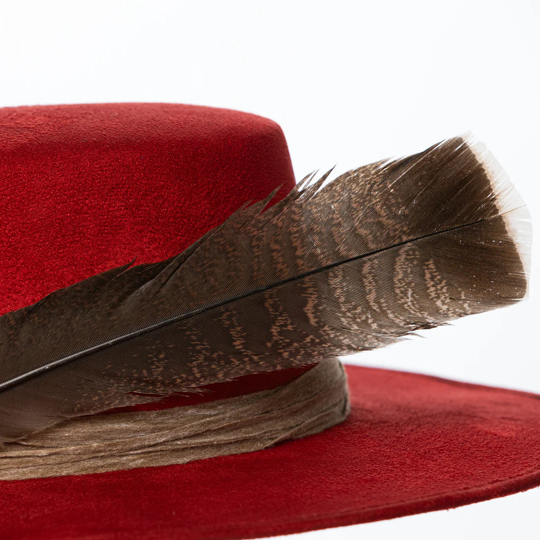 Close-up of brown feather resting on layered rustic band of red suede hat, highlighting artisanal detail and natural elements.
