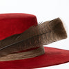 Close-up of brown feather resting on layered rustic band of red suede hat, highlighting artisanal detail and natural elements.