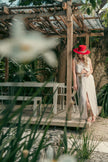 Blonde model wearing the red feather hat and beige linen set, standing barefoot in a lush Caribbean patio with rustic pergola.