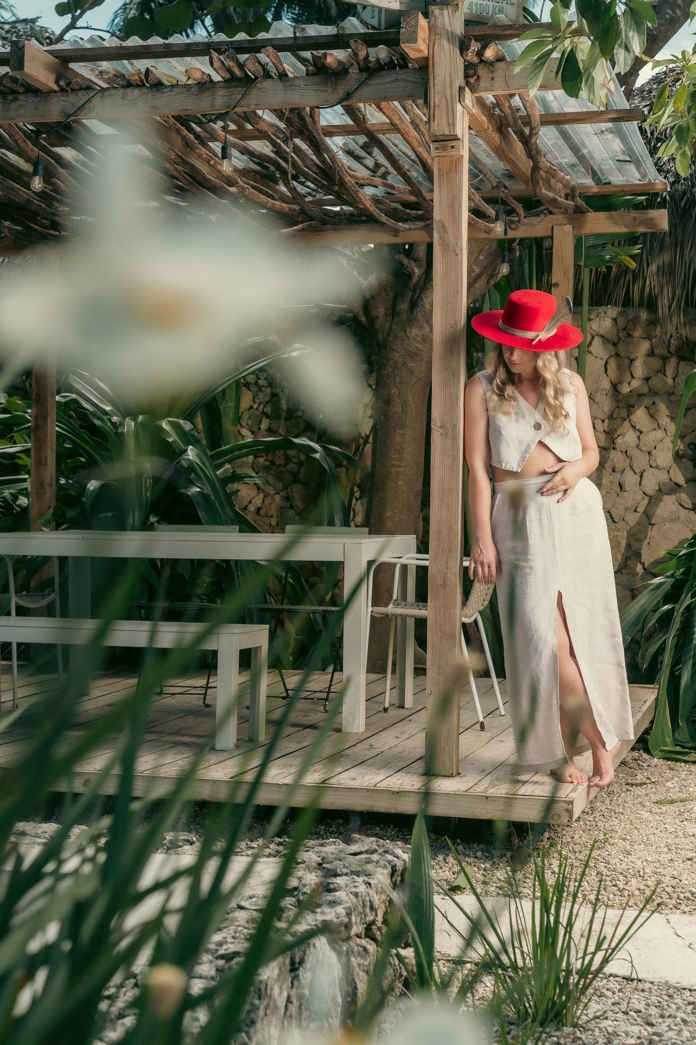 Blonde model wearing the red feather hat and beige linen set, standing barefoot in a lush Caribbean patio with rustic pergola.