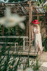 Blonde model wearing the red feather hat and beige linen set, standing barefoot in a lush Caribbean patio with rustic pergola.