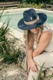 Blonde model wearing the See You in My Dreams hat, seated near a pool in a tropical garden, exuding serene elegance.